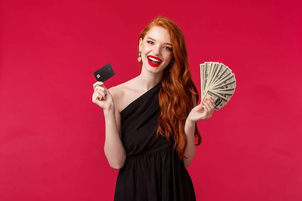Luxury, beauty and money concept. Elegant good-looking redhead woman being rich, wear luxurious black dress, holding credit card and fan of dollars, brag her income, smiling carefree