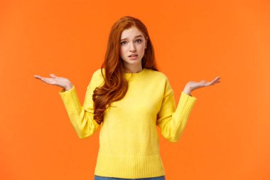 Şaşkın kızıl saçlı kız, omuz silkiyor ve kafası karışmış bakıyor, cevap vermiyor, yardımcı olamıyor, kafası karışık ya da kararsız, anlayamıyorum, turuncu arka planda duruyor.