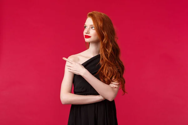 Celebración, eventos, concepto de moda. Retrato de la joven pelirroja elegante mujer delgada en vestido elegante negro, usar maquillaje lápiz labial rojo, mirar y señalar a la izquierda con la cara interesada curiosa — Foto de Stock