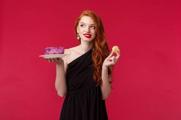 Pensativo e sonhador linda mulher ruiva magro em vestido preto pensando em comer sobremesa enquanto cuida de seu corpo, desviar o olhar ponderando e sorrindo, segurar biscoito e bolo — Fotografia de Stock