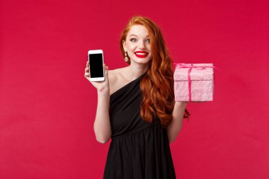 Heyecanlı ve neşeli gülümseyen kızıl saçlı kadının portresi bu kadar çabuk hediye almamış, cep telefonu uygulamasını, online dağıtım uygulamasını, pembe hediyeyi kırmızı arkaplan üzerinde tutmamış.