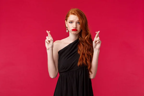 Retrato de esperanzadora pelirroja mujer caucásica preocupada en vestido de lujo negro, dedos cruzados buena suerte, quieren ganar, haciendo que el deseo mueca nervioso, inseguro en sus propias posibilidades, fondo rojo — Foto de Stock