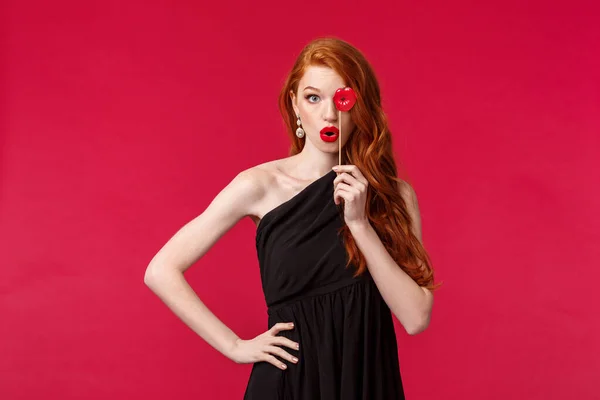 Concepto de celebración, fiestas y mujeres. Retrato de mujer pelirroja joven descarada y femenina en vestido negro con estilo, la celebración de los labios tarjeta sobre el ojo y plegable propia en signo de beso mwah, fondo rojo —  Fotos de Stock