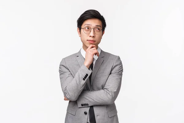 Waist-up portrait of smart and creative young asian male entrepreneur in grey suit, making plan, hold hand on chin and looking up thoughtful, thinking over project, standing white background