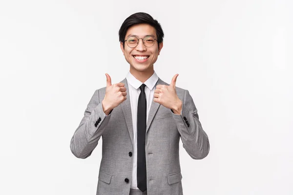 Waist-up portrait of pleased, fully satisfied asian young man in grey suit, showing thumbs-up and smiling with approval, like awesome concept. recommend service, assure it is best deal