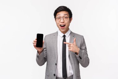 Şaşırmış ve heyecanlı Asyalı genç ofis çalışanının belden yukarı portresi akıllı telefon ekranında harika bir şey gösteriyor, cep telefonunu tutuyor ve etkileyici bir yüz ifadesiyle görüntüyü işaret ediyor.