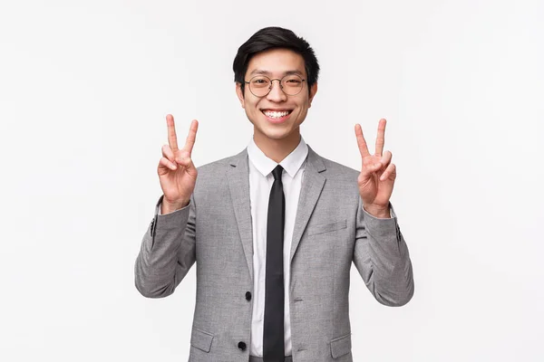 Midja upp porträtt av optimistisk glad och vacker asiatisk man i grå kostym, glasögon som visar fred tecken, leende kawaii, ta löjlig bild, känna sig glad och glad, vit bakgrund — Stockfoto