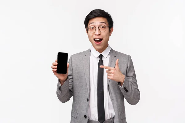 Cintura-up retrato de espantado e animado asiático jovem trabalhador de escritório mostrando algo incrível na tela do smartphone, segure o telefone celular e apontando para a tela com o rosto impressionado — Fotografia de Stock