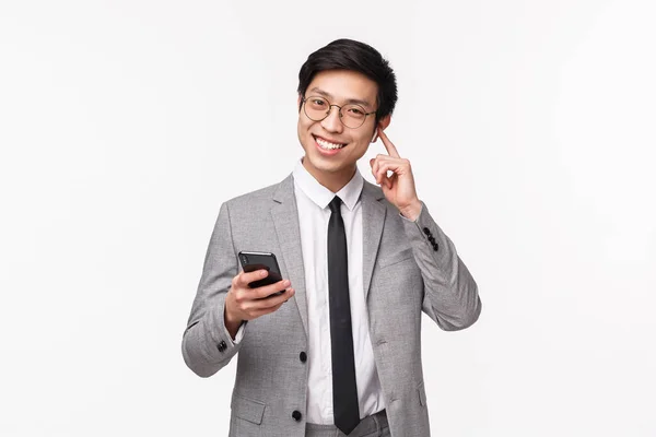 Retrato en la cintura de un feliz hombre de negocios asiático satisfecho y sonriente con auriculares inalámbricos, sosteniendo el teléfono móvil, cambiando el volumen o llamando a alguien con auriculares, de pie fondo blanco — Foto de Stock