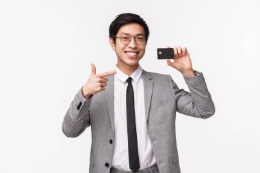 Tatmin olmuş, yakışıklı, takım elbiseli, emin Asyalı bir adamın belden yukarı portresi, kredi kartı gösteriyor, mutlu bir gülümsemeyle işaret ediyor, nakit olmayan ödemeler yapmayı öneriyor, beyaz bir geçmişi var.