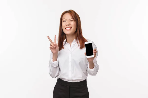 Porträt einer fröhlichen, gut aussehenden Asiatin in weißem Hemd, Rock, Augenzwinkern und zufriedenem Lächeln, mit Peace-Kawaii-Zeichen als Smartphone-Applikation, Handy-Display, weißem Hintergrund — Stockfoto