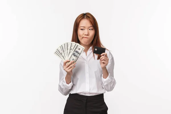 Porträtt av tveksam och tveksam söt asiatisk kvinna köper dyra sak, tänker betala med kreditkort eller kontanter, tittar på pengar funderar och vara osäker, stå vit bakgrund — Stockfoto