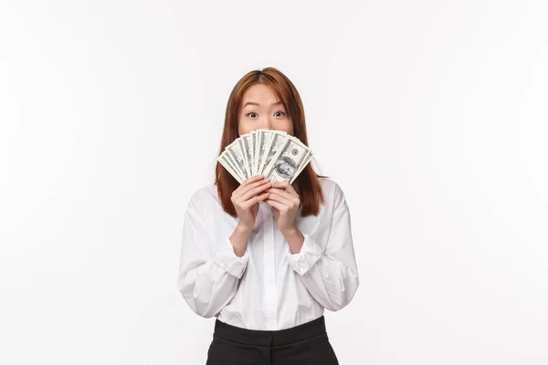 Portrait of excited and pleased asian woman got rich, telling secret of wealth, hiding face behind money, holding fan of cash and smiling with eyes at camera, stand white background — Stock Photo, Image