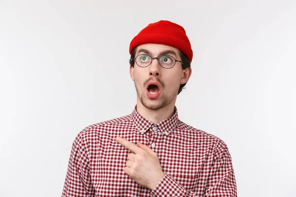 Primer plano retrato sorprendido y sin palabras divertido hombre caucásico con barba, en gafas y gorro rojo, caída de la mandíbula mirada y señalando esquina superior izquierda asombrado, ver algo impresionante, fondo blanco — Foto de Stock