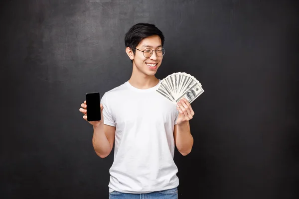 Concepto de tecnología, dinero y premios. Agradable sonriente feliz rico asiático chico en camiseta, mirando satisfecho en dólares fan de dinero en efectivo y la celebración de teléfono móvil, mostrando la pantalla del teléfono inteligente, fondo negro — Foto de Stock