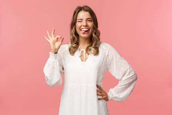 Retrato de feliz sorrindo mulher loira com corte de cabelo encaracolado curto, mostrar a língua e sinal ok, dizer tudo bem, concordar ou aprovar, taxa grande produto, garantia de qualidade, stand pink background — Fotografia de Stock