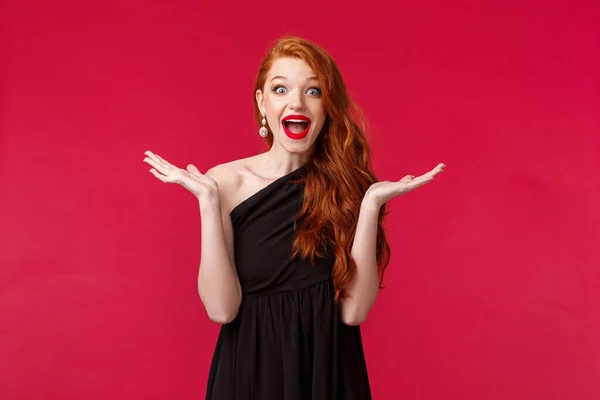 Fashion, luxury and beauty concept. Portrait of happy and surprised excited redhead woman react to unexpected pleasant guest showing-up her party, raise hands sideways and smiling, black dress