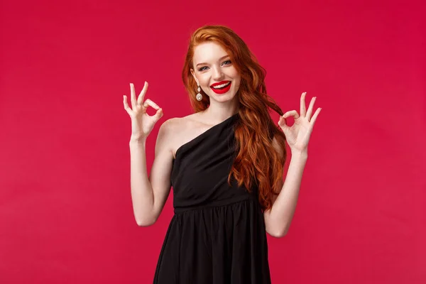 Concepto de moda, lujo y belleza. Retrato de mujer pelirroja sensual muy guapa en vestido negro y maquillaje, mostrando bien, excelente o signo de garantía, fondo rojo — Foto de Stock