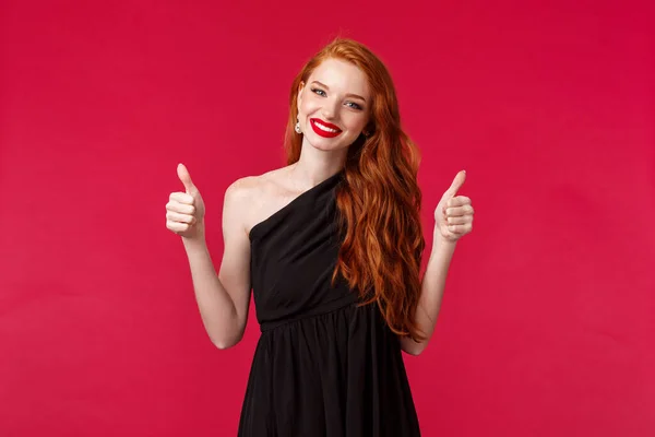 Mode, luxe et concept beauté. Portrait de charmante jeune femme rousse de soutien en robe élégante noire, maquillage, sourire heureux montrer pouces vers le haut dans l'approbation ou comme, fond rouge — Photo