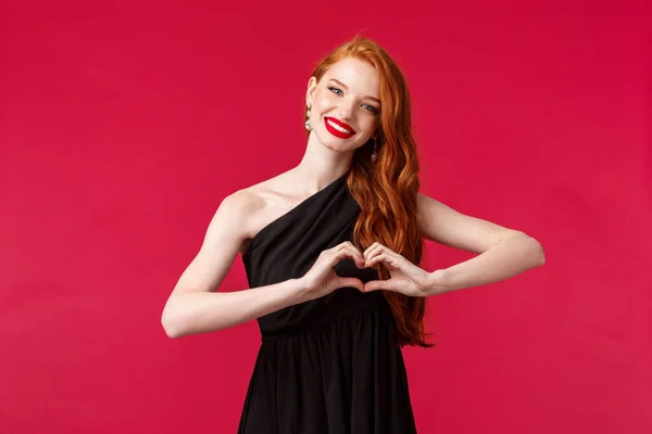 Retrato de encantadora chica caucásica elegante en lápiz labial rojo, vestido negro con pelo largo rojo, mostrando el signo del corazón sobre el pecho y el corazón sonriente reconfortante, expresar simpatía y aprecio, fondo rojo — Foto de Stock