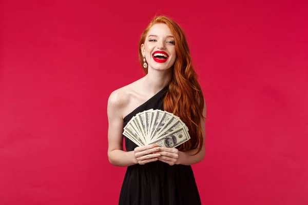 Luxury, beauty and money concept. Girl got expensive taste. Happy rich good-looking redhead woman in stylish black dress, laughing delighted, holding dollars, congratulate winner with prize