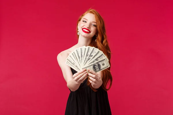 Concepto de lujo, belleza y dinero. Retrato de hermosa sensual joven pelirroja en vestido negro felicitar al afortunado ganador con el premio, dando dólares recompensa en efectivo, cámara sonriente, fondo rojo — Foto de Stock