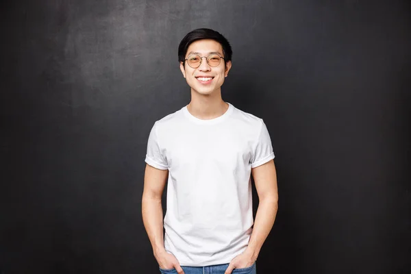 Портрет молодого азиатского парня в очках, стоящего в белой повседневной рубашке на черном фоне, дружелюбно улыбающегося, выражающего радостные эмоции, зависающего с друзьями после колледжа — стоковое фото
