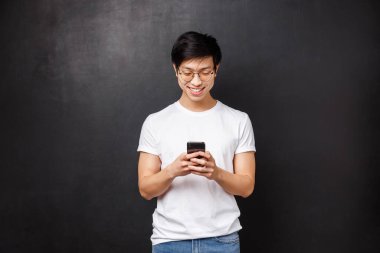 Teknoloji, aletler ve insan konsepti. Telefonla oynayan gülümseyen Asyalı genç adam, akıllı telefon mesajcısını kullanarak arkadaşına mesaj atıyor, sosyal medyayı düzenlemek için yeni bir uygulama kullanıyor, siyah arka plan,
