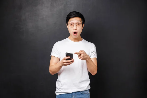 Technology, gadgets and people concept. Impressed and speechless fascinated asian guy saw something super cool in internet, telling about it at camera, open mouth say wow, hold mobile phone