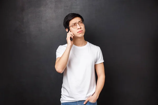 Technology, gadgets and people concept. Portrait of thoughtful young asian guy thinking over question, calling friend, talking on phone, hold smartphone and look away pondering what answer — Stock Photo, Image