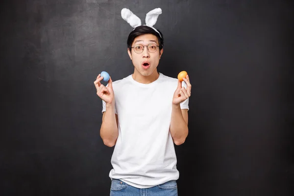 Feiertage, Party und Ostern. Erstaunte junge asiatische Kerl in Hasenohren mit bemalten Eiern als Lernen Freundin Traditionen, offener Mund fasziniert neue Kultur kennen zu lernen, schwarzer Hintergrund — Stockfoto