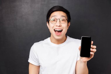 Teknoloji, mesajlaşma ve insan konsepti. Beyaz tişörtlü mutlu, şaşırmış ve etkilenmiş Asyalı genç adamın yakın plan portresi, akıllı telefon görüntüsü sergiliyor.