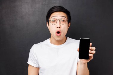 Teknoloji, mesajlaşma ve insan konsepti. Şaşırmış ve heyecanlı Asyalı genç adamın yakın plan portresi, cep telefonu ekranını gösteriyor, fantastik yeni uygulama hakkında konuşuyor, siyah arkaplan