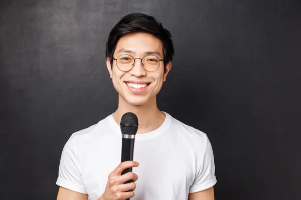 Leisure, people and music concept. Handsome and cute smiling asian man in white t-shirt, glasses, holding microphone, singing at karaoke party with friends, picking song on screen, black background