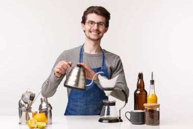 Barmen, kafe çalışanı ve barmen konsepti. Önlüklü neşeli görünümlü genç sakallı adamın portresi çaydanlıktan su dökerken müşteriye gülümsüyor ve filtre kahve yapıyor.