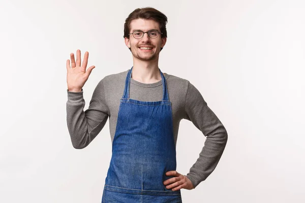 Taille-up portret van vriendelijke uitgaande jonge mannelijke barista in schort, gedag zeggen tegen de klant, mensen in cafe begroeten als zwaaiend hand hallo teken, vrolijk glimlachen, orde op zaken stellen koffie of drinken — Stockfoto