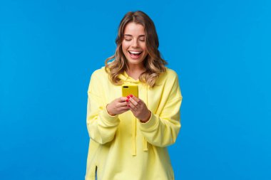 Teknoloji ve insan konsepti. Sarı çantada cep telefonu kullanan mutlu, gülümseyen sarışın kız, mesajlaşan akıllı telefon görüntüsüne mutlu bir ifadeyle bak, komik mim gönder, mavi arka plan