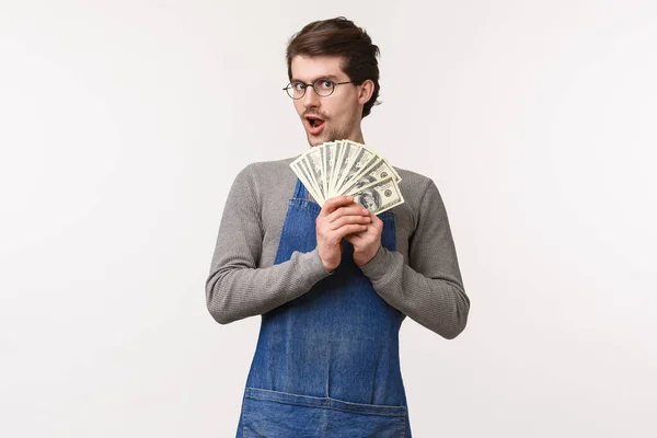 Concepto de pequeña empresa, finanzas y carrera. Entusiasta joven estudiante masculino que trabaja a tiempo parcial en la cafetería, usa delantal sosteniendo su salario, fan de los dólares, ganar dinero, fondo blanco — Foto de Stock