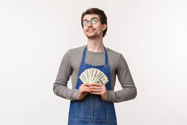 Concepto de pequeña empresa, finanzas y carrera. Retrato de joven soñador y complacido, guapo en delantal, trabajando en una cafetería o restaurante en delantal, reteniendo dinero y pensando cómo desperdiciarlos — Foto de Stock