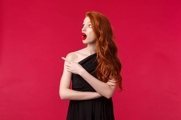 Celebration, events, fashion concept. Portrait of amazed and shocked redhead young pretty woman in elegant black evening dress, drop jaw, looking and pointing left astonished, red background — 스톡 사진