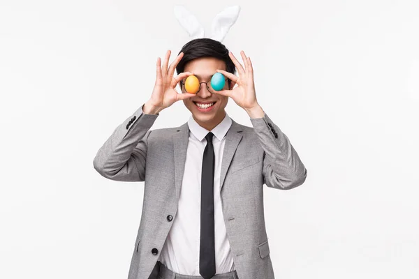 Vacances, personnes et concept de célébration. Portrait taille haute d'un gars asiatique drôle, ludique et enthousiaste en costume gris et cravate, tenant des œufs peints sur les yeux et souriant, profiter de la fête de Pâques — Photo