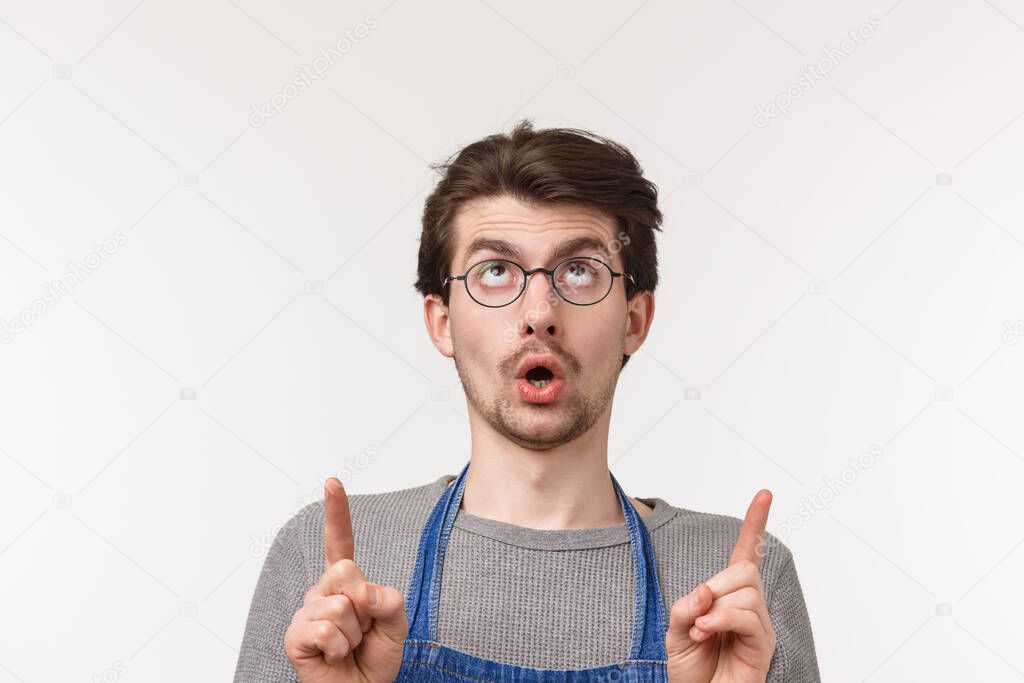Advertisement, small business and career concept. Close-up portrait of curious and intrigued caucasian man in apron and glasses spot something interesting upwards, pointing fingers up