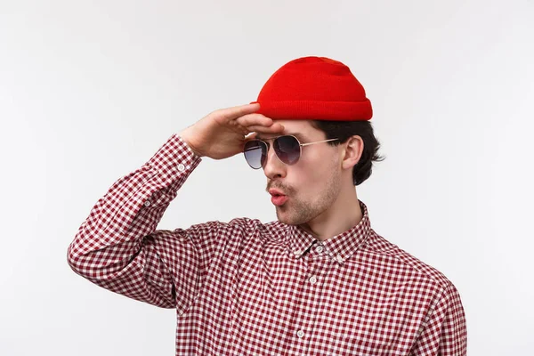 Close-up retrato de jovem hipster intrigado e animado em disfarce, óculos de sol e gorro vermelho, olhando para a distância, ver algo super legal longe, ficar espantado sobre fundo branco — Fotografia de Stock