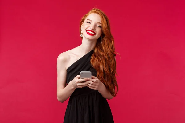 Portrait of gorgeous smiling woman with ginger hair in elegant black prom dress, holding smartphone, messaging, using application, want take picture, laughing and looking camera, red background — Stock Photo, Image