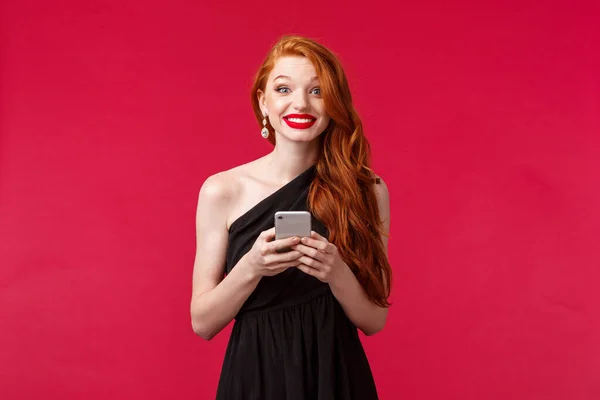Portrét vzrušené a pobavené, zrzka žena v černých šatech na její rande nebo ples noc, držení mobilního telefonu, úsměv zářící na kameru, stojící červené pozadí pozitivní — Stock fotografie