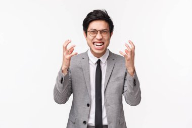 Gri takım elbiseli nefret dolu, öfkeli Asyalı bir adamın belden aşağılayıcı portresi yumruklarını sıkması, suratını buruşturması ve kameraya karşı öfkeyle bakması, beyaz arka planda durması.