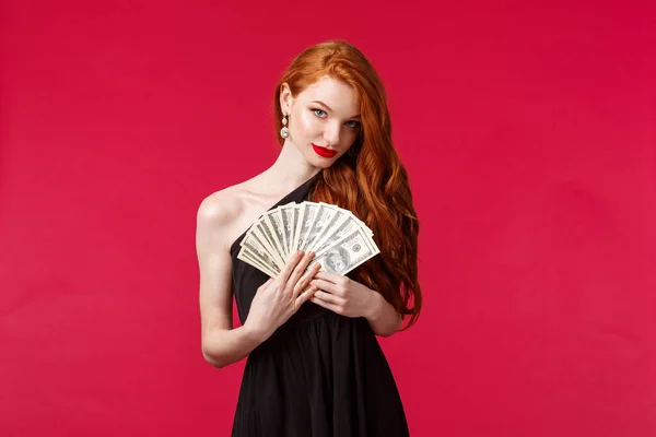 Luxury, beauty and money concept. Portrait of sensual good-looking, flirty young redhead woman in black stylish dress, hinting you can get this cash dollars if place good bet, smiling daring