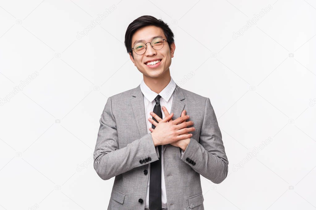 Waist-up portrait of grateful, flattered asian young male office manager, entrepreneur hold hands on heart and smiling, thanking for compliment, flattered and grateful for praises, white background
