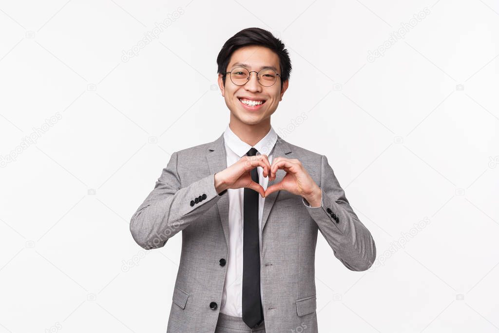 Waist-up portrait of lovely, cute asian young man, office manager in suit, showing heart sign and smiling blushing, expressing his sympathy, confessing in love, standing white background