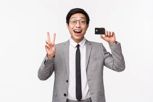 Ένα πορτραίτο ενθουσιώδους νεαρού Ασιάτη επιχειρηματία με κοστούμι, με πιστωτική κάρτα και σήμα ειρήνης, χαμογελαστό χαρούμενο, εντυπωσιασμένο με την επιστροφή χρημάτων και νέα τραπεζικά χαρακτηριστικά για τους πελάτες — Φωτογραφία Αρχείου